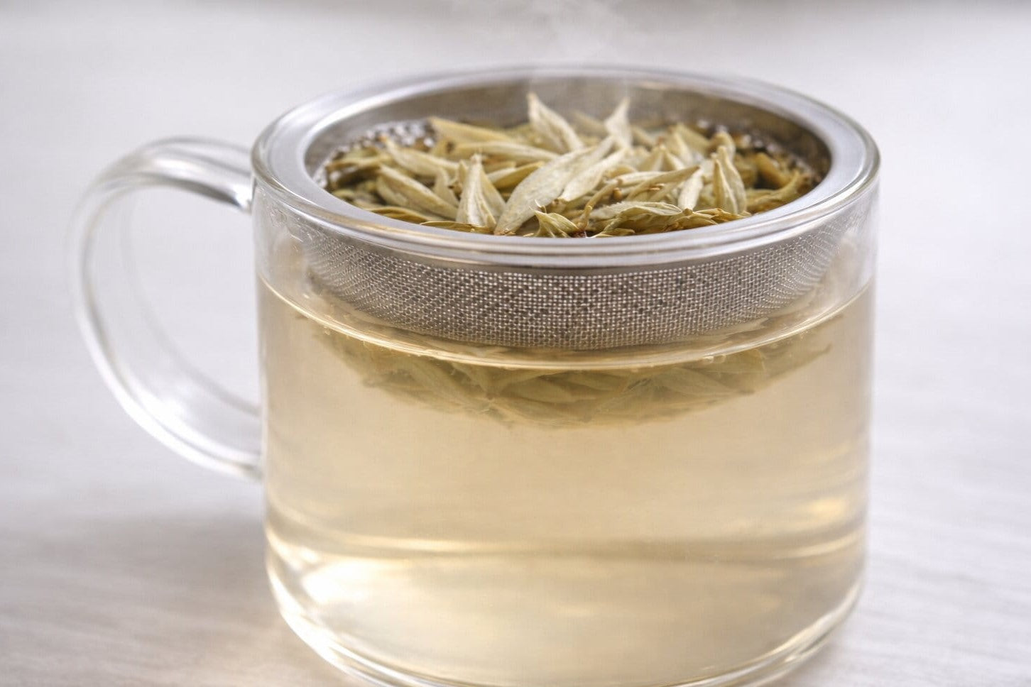 Clear glass mug with a stainless steel tea infuser containing white tea leaves steeping in hot water, light golden liquor with subtle steam.